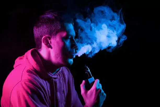 Close up portrait of the face of an adult serious man exhales blue toxic smoke while smoking e-cigarette and vape illuminated with pink colored light on a black background. Harm to health.